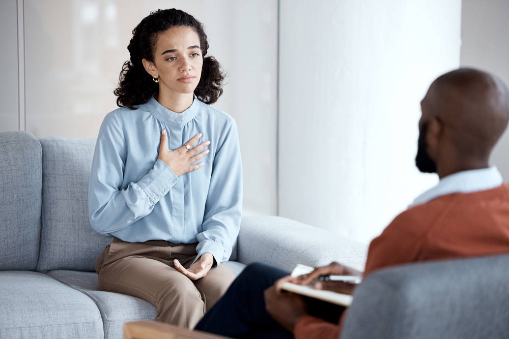 A young woman sits in a therapist's office with her hand on her chest.