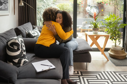 A teen girl sits in a therapists office, hugging another woman.